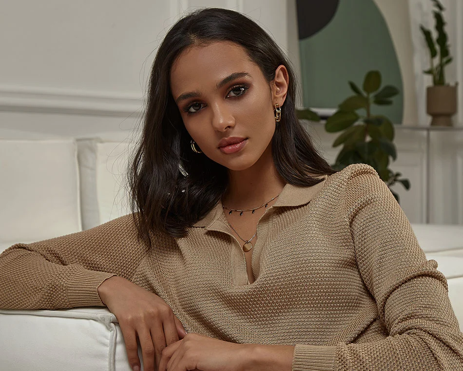 A woman with dark hair wearing a beige sweater, seated comfortably on a sofa, looking into the camera with a confident and relaxed expression - Botox in Grand Rapids, MI