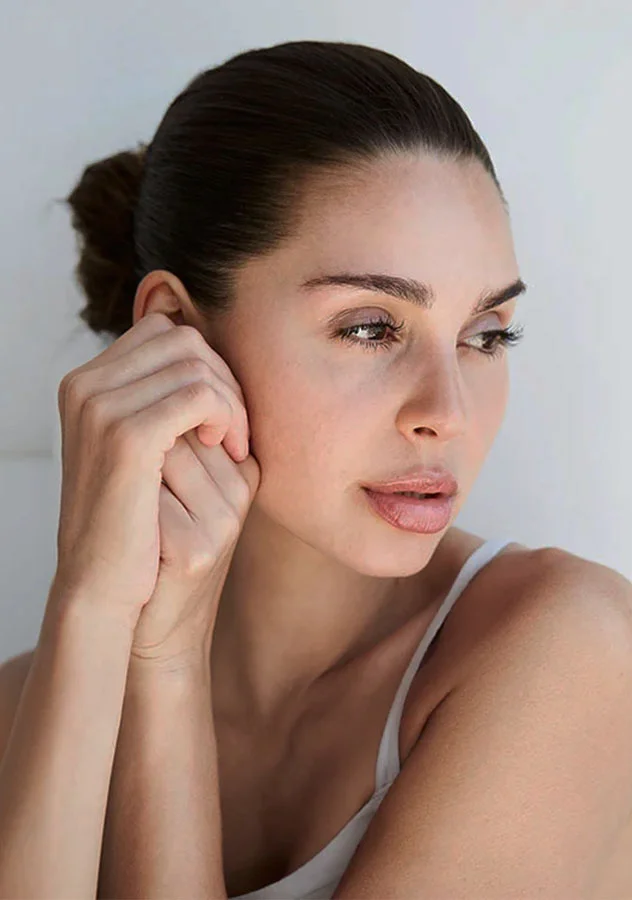 A woman with her hair tied back, gently touching her cheek, showcasing her smooth, glowing skin in soft lighting - Botox in Grand Rapids, MI