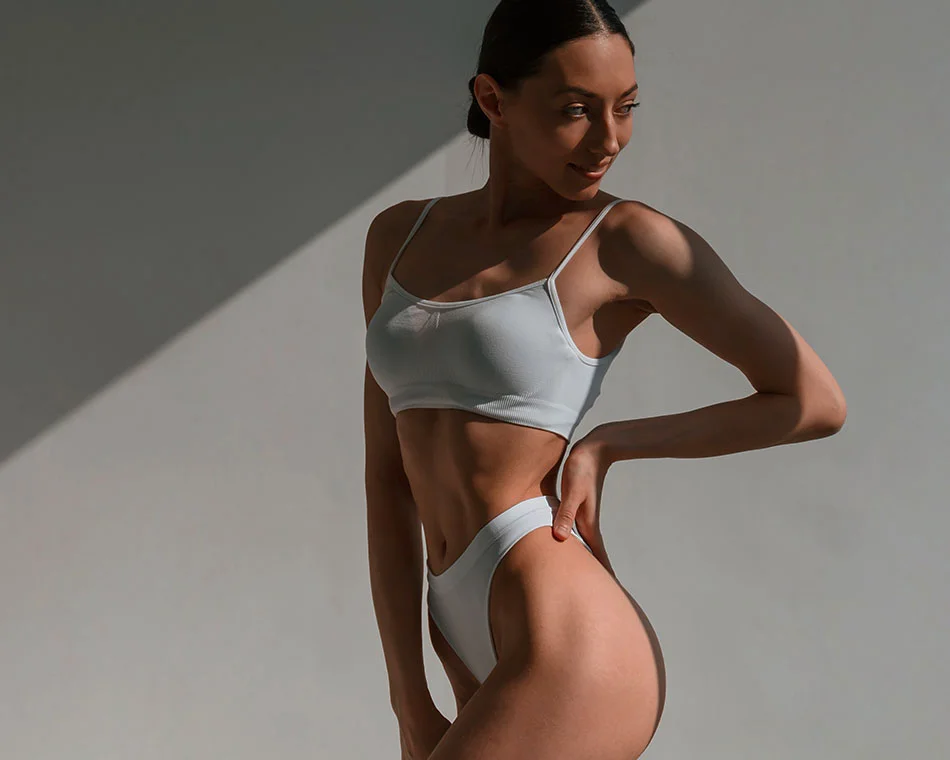 Fit woman posing in a minimal studio setting, showcasing her toned physique in a light gray sports bra and high-cut bottoms, highlighting body confidence and fitness - Brazilian Butt Lift in Grand Rapids, MI