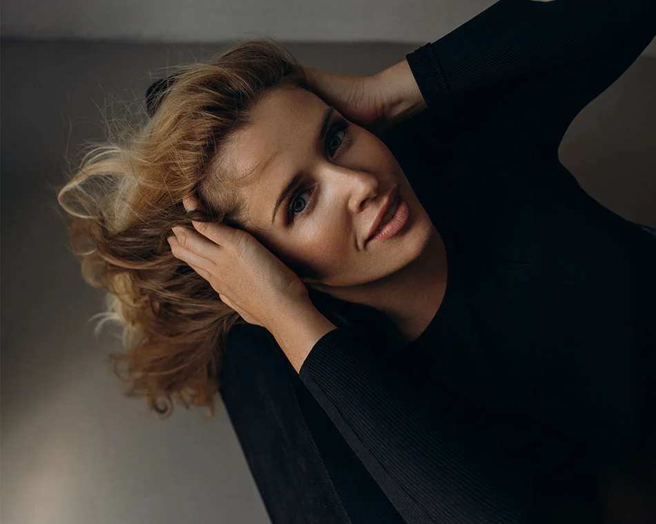 a woman reclining with her hands resting on her head. She has a confident and relaxed expression, with her blonde hair cascading around her - Brow Lift in Grand Rapids, MI