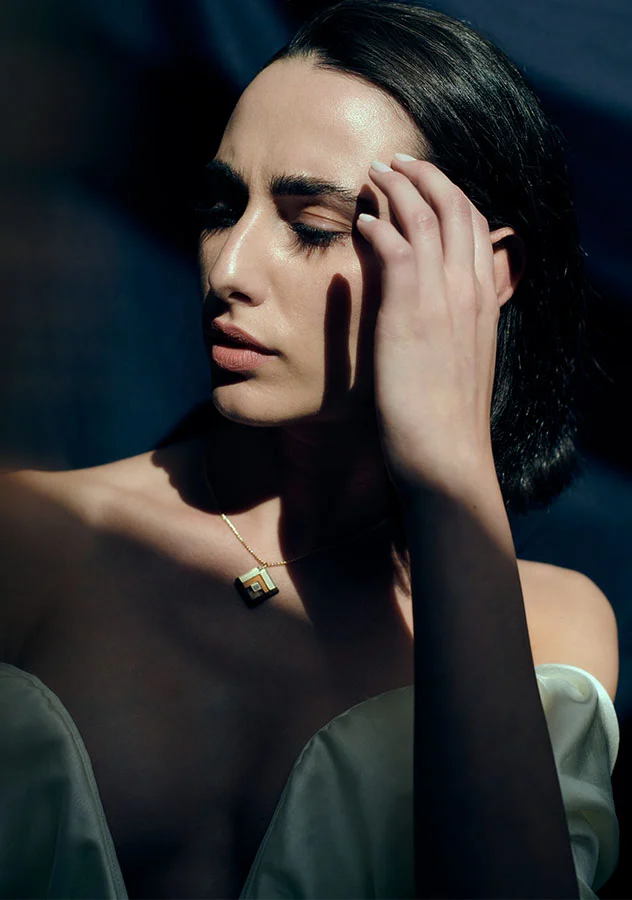 a woman with dark, slicked-back hair, touching her face in a thoughtful pose. She is illuminated by natural light, creating soft shadows that highlight her features - Brow Lift in Grand Rapids, MI