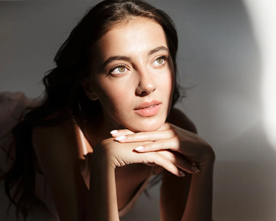 A woman with long brown hair, resting her chin on her hands, gazes thoughtfully into the distance with natural light accentuating her facial features - Chin Augmentation in Grand Rapids, MI