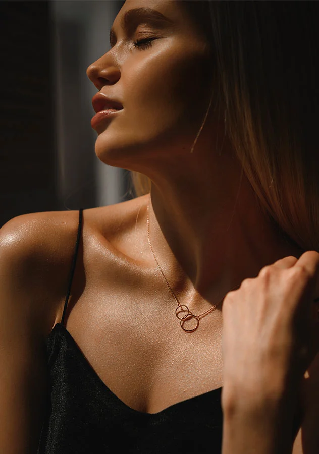 A woman with her eyes closed, wearing a black strappy top and a delicate necklace, standing in dramatic lighting that emphasizes her glowing skin - Clear and Brilliant Treatment in Grand Rapids, MI