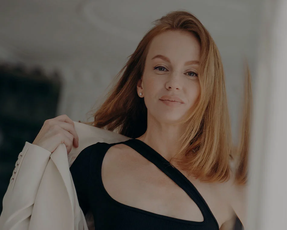 Close-up of a woman with red hair, smiling confidently while wearing a black outfit and white blazer - Facial Fat Grafting in Grand Rapid, MI