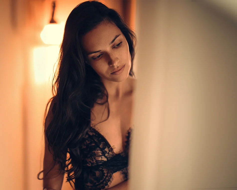 A woman with long hair, dressed in black lace, looking down thoughtfully in a dimly lit room - Male to Female in Grand Rapids, MI