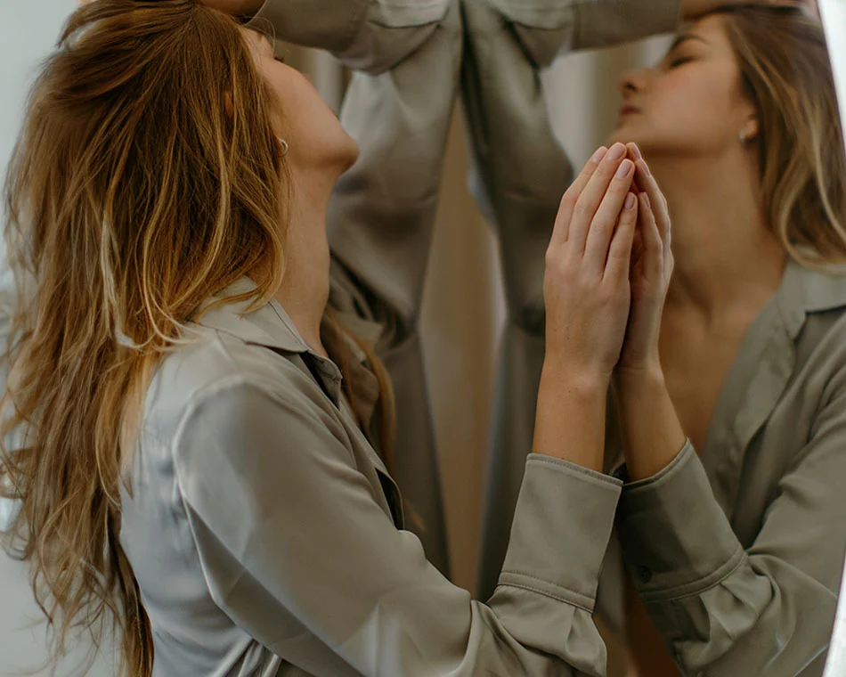 A woman standing in front of a mirror, with her head tilted back, hands pressed against the glass as she closes her eyes, wearing a silk shirt - Neck Lift in Grand Rapids, MI