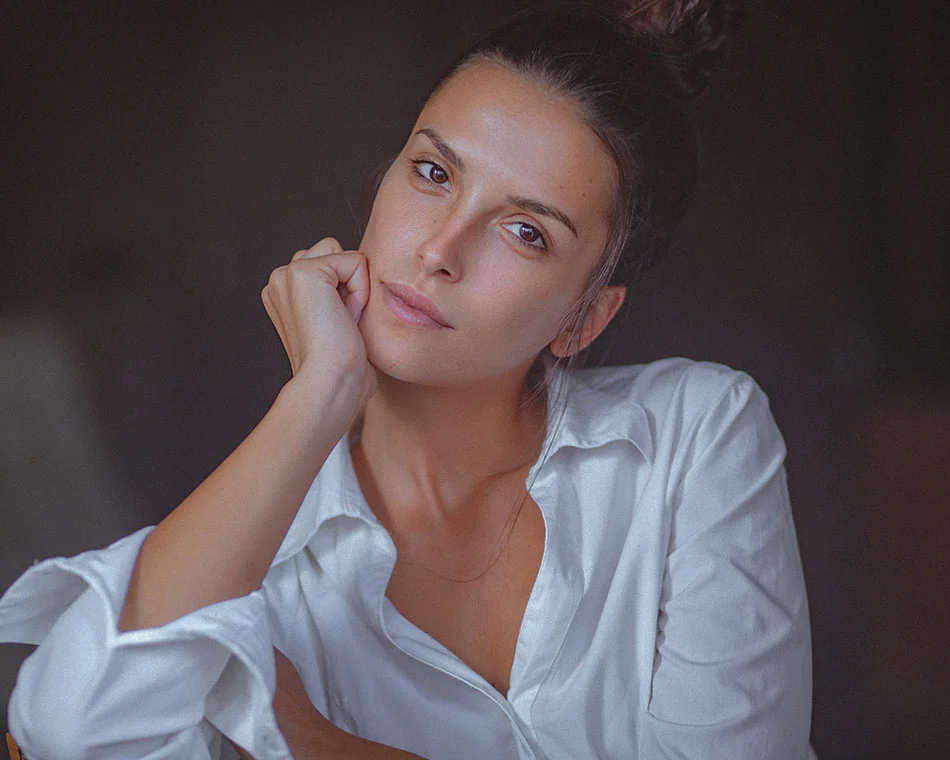 Casual portrait of a woman with her hair in a bun, wearing a white shirt and resting her chin on her hand, conveying a relaxed and thoughtful expression - Sculptra in Grand Rapids, MI