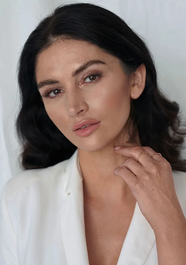 Close-up of a woman with dark wavy hair, wearing a white outfit and lightly touching her neck, showcasing natural beauty and elegance - Sculptra in Grand Rapids, MI